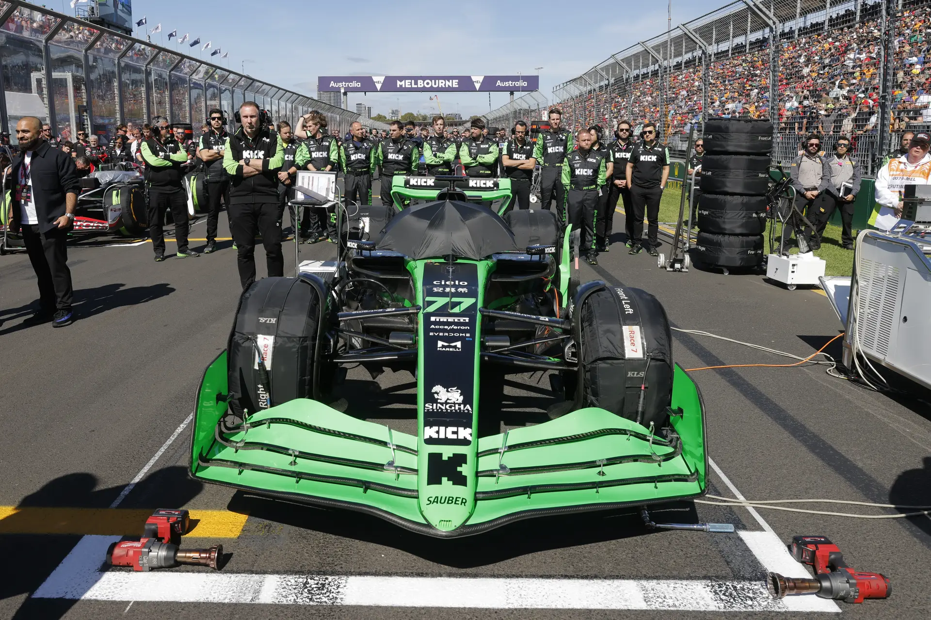 A green and black Formula 1 car positioned on the starting grid, surrounded by team members and crew, with a packed grandstand in the background.