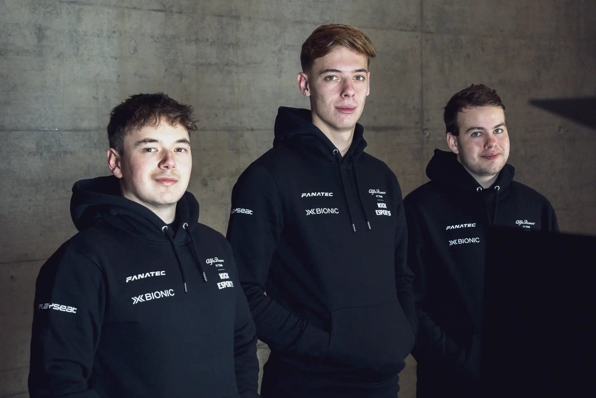 Three Sauber Esports drivers posing in team jackets against a dark concrete background.
