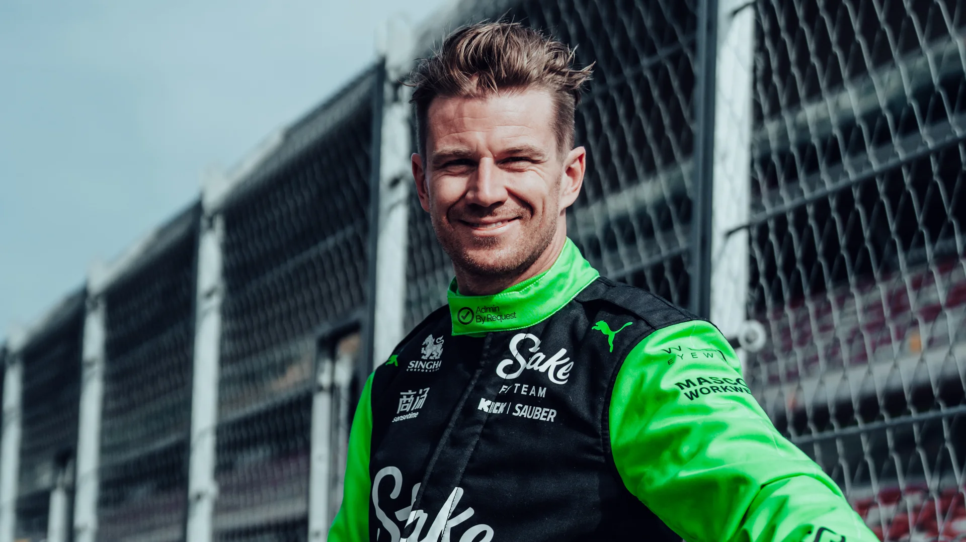 Nico Hülkenberg at TV Day in Barcelona, wearing his green Sauber F1 racing suit, smiling at the camera with the racetrack in the background.