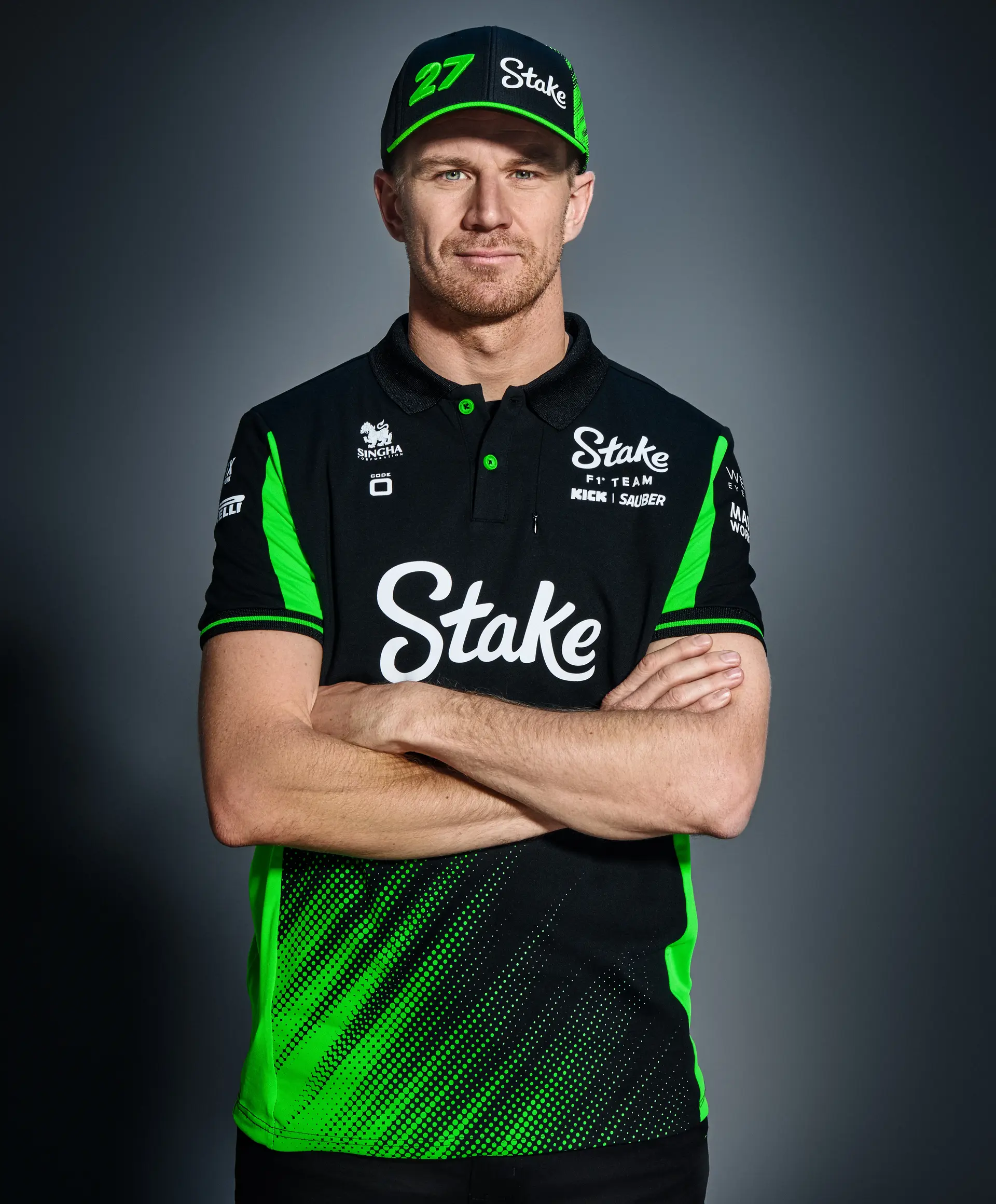 F1 driver Nico Hülkenberg posing with arms crossed, wearing the Sauber F1 Team's 2025 Code-Zero teamwear in black and green, featuring Stake sponsorship branding and a matching cap.