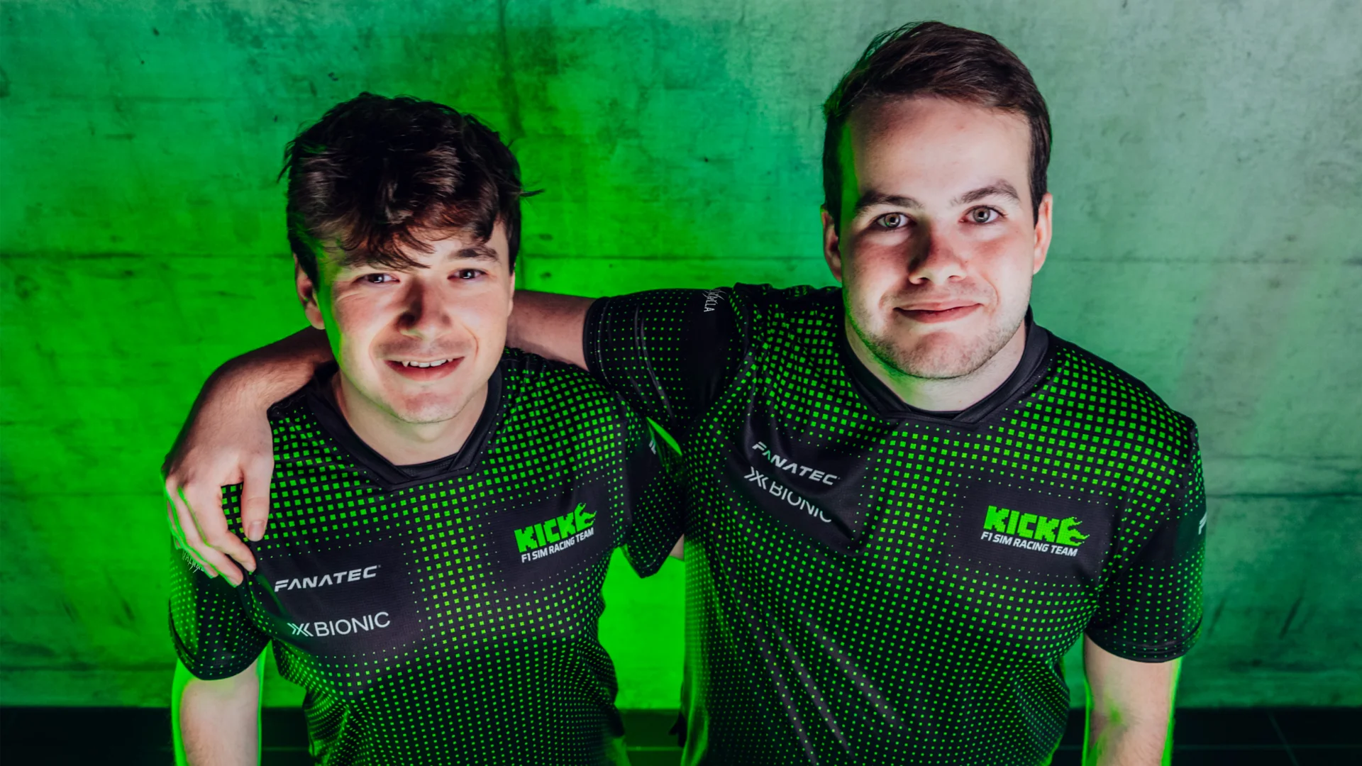 Two members of the KICK F1 Sim Racing Team posing in their newly launched 2025 team kit, featuring a vibrant green and black design under neon lighting.