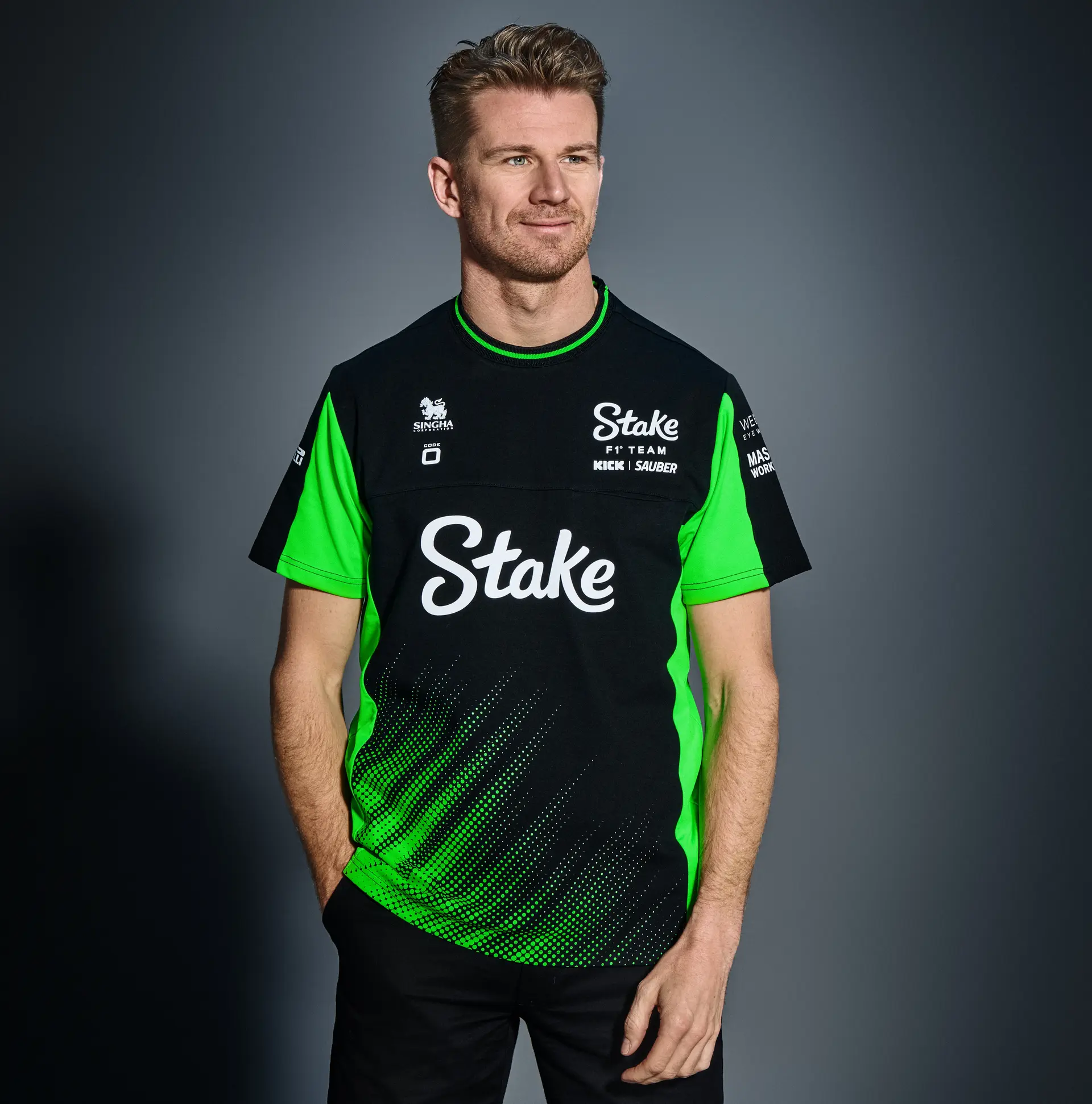 F1 driver Nico Hülkenberg wearing the Sauber F1 Team's 2025 Code-Zero teamwear, featuring a black and green gradient design with Stake branding, posing with his hands in front.