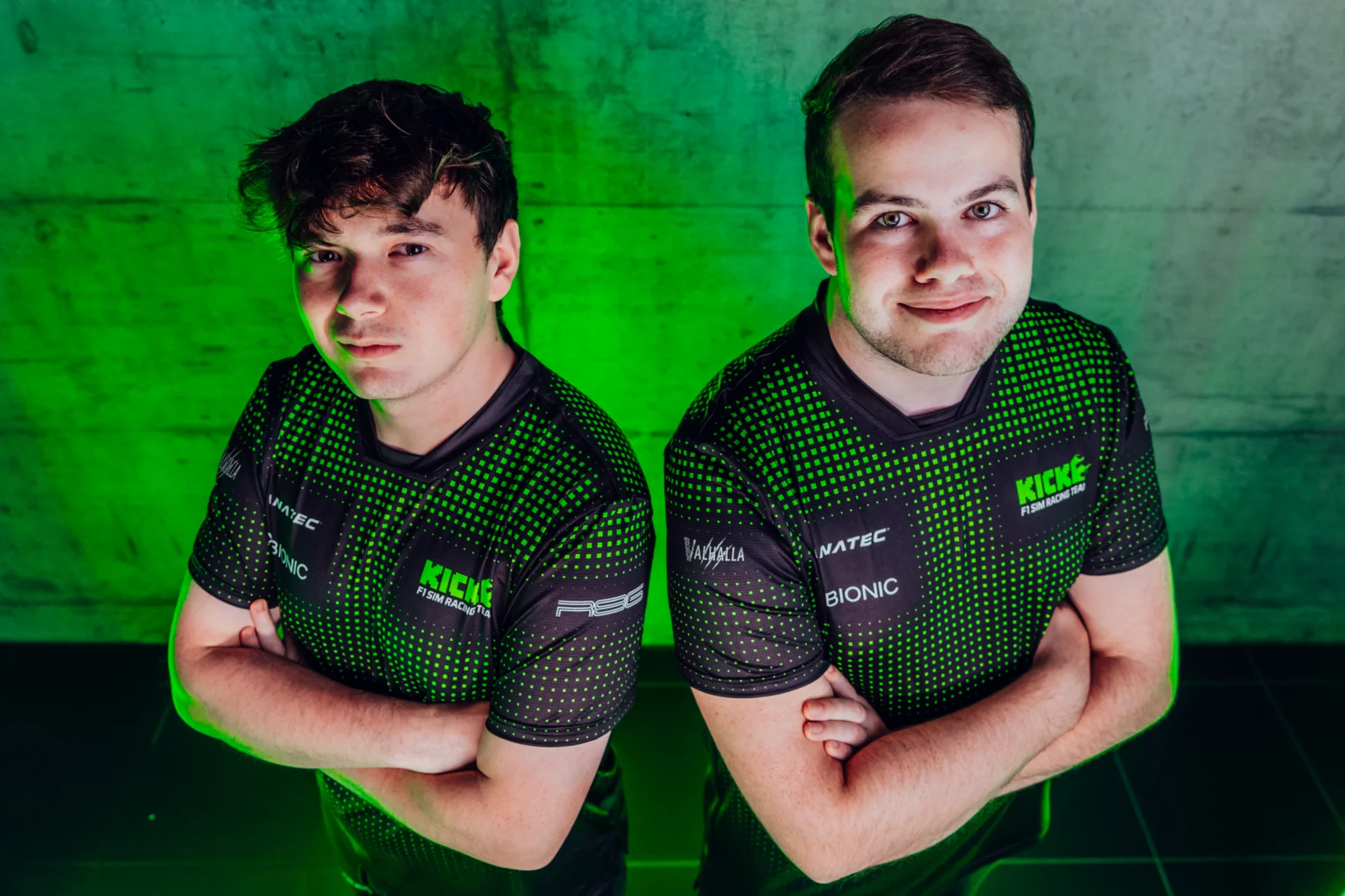 Two KICK F1 Sim Racing Team drivers wearing green and black team jerseys stand against a green-lit background, highlighting the team's partnership with the Floki-powered Valhalla platform to enhance fan engagement.